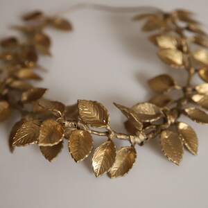 Może przedstawiać: Złota korona z liści o prostym wzorze. Korona wykonana jest z pojedynczych liści połączonych ze sobą w okrąg.