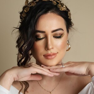 Może przedstawiać: Kobieta o ciemnych włosach, w złotej koronie z liści, złotym cieniu do powiek i delikatnym złotym naszyjniku. Ma na sobie białą koszulę i trzyma ręce na brodzie.