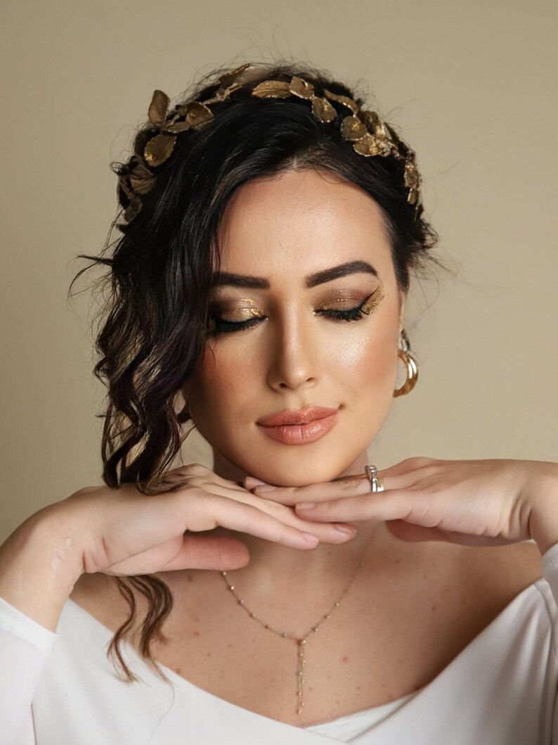 Może przedstawiać: Kobieta o ciemnych włosach, w złotej koronie z liści, złotym cieniu do powiek i delikatnym złotym naszyjniku. Ma na sobie białą koszulę i trzyma ręce na brodzie.