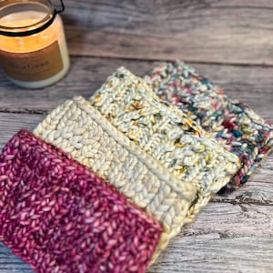 May include: Four chunky knit headbands in shades of pink, cream, and multicolored yarn. The headbands are stacked on a wooden surface, with a lit candle in a glass jar in the background. The headbands have a button detail.
