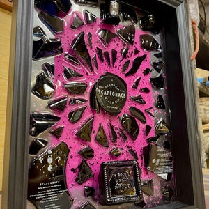 May include: A framed artwork featuring a black bottle of Scapegrace Black Gin, surrounded by broken black glass pieces embedded in a pink resin background. The bottle is labeled "Scapegrace Black Gin" and "Premium Small Batch Gin".
