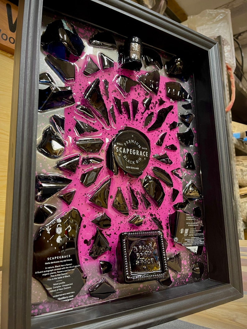 May include: A framed artwork featuring a black bottle of Scapegrace Black Gin, surrounded by broken black glass pieces embedded in a pink resin background. The bottle is labeled "Scapegrace Black Gin" and "Premium Small Batch Gin".
