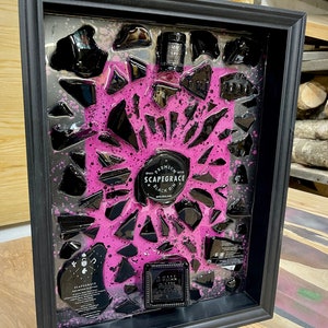 May include: A framed artwork featuring a black and pink design with broken glass pieces. The artwork includes a bottle of "Scapegrace Black Gin" and a label that reads "Small Batch Premium New Zealand Gin".
