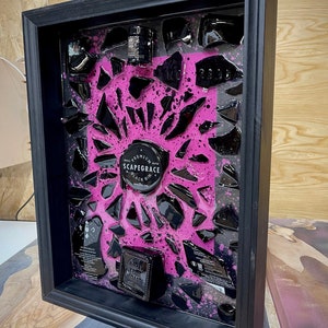 May include: A framed artwork featuring a black bottle of Scapegrace Black Gin, broken into pieces and encased in pink resin. The bottle is surrounded by black shards and pink splatter paint.