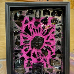 May include: A black framed artwork with a pink background and black glass shards. The artwork features a small bottle of "Scapegrace Black Gin" and a label with the text "Small Batch Premium Scapegrace Black Gin New Zealand".
