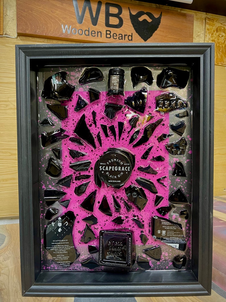 May include: A black framed artwork with a pink background and black glass shards. The artwork features a small bottle of "Scapegrace Black Gin" and a label with the text "Small Batch Premium Scapegrace Black Gin New Zealand".