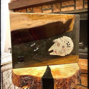 May include: A wooden art piece featuring a Star Wars theme. The artwork depicts the Millennium Falcon and TIE fighters against a dark, space-like background. The piece is set on a rustic, wooden table.