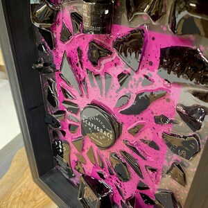 May include: A framed piece of art featuring a black and pink abstract design with a small bottle and a round label that reads "Scapegrace Black Gin, New Zealand".