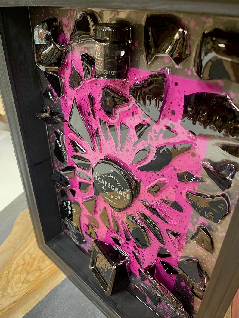 May include: A framed piece of art featuring a black and pink abstract design with a small bottle and a round label that reads "Scapegrace Black Gin, New Zealand".
