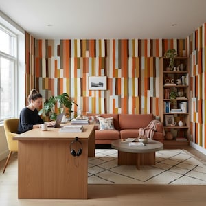 Op de afbeelding: Een thuiskantoor met een houten bureau, een gele stoel en een roestkleurige bank. De kamer heeft een behang met patronen in tinten oranje, bruin en wit. Een vrouw werkt op een laptop aan het bureau. Een boekenkast en een ronde salontafel maken de ruimte compleet.