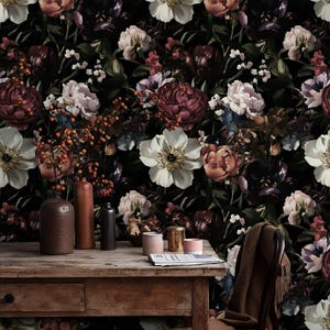 Op de afbeelding: Donker bloemenbehang met grote witte, perzik- en bordeauxrode bloemen op een zwarte achtergrond. Een houten tafel toont vazen en kleine containers, wat een vintage esthetiek creëert. Het ontwerp straalt klassieke elegantie uit.