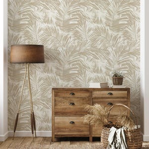 Pode incluir: Interior de um quarto com uma cômoda de madeira, um candeeiro de pé e um cesto de vime. A parede é coberta com papel de parede com um padrão repetido de folhas de palmeira em bege e branco sujo. Um tapete cobre o chão de madeira.