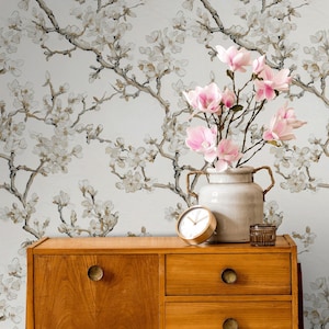 May include: A wooden cabinet with two drawers and a door, topped with a vase of pink flowers, a gold clock, and a small glass jar. The background is a floral wallpaper with branches and white flowers.
