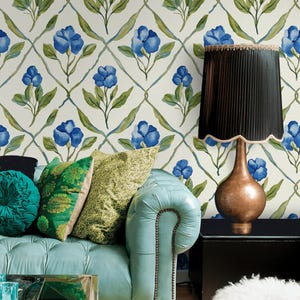 May include: A living room scene featuring a light blue leather sofa with decorative pillows in shades of green. The background showcases floral wallpaper with blue flowers and green leaves. A copper-toned lamp with a black shade sits on a black side table.