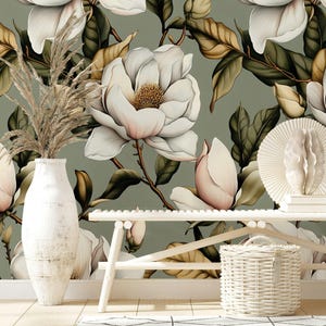 Puede incluir: Un diseño de papel pintado floral con grandes flores de magnolia blancas con acentos rosas y hojas verdes sobre un fondo verde apagado. Un jarrón blanco con hierba de la pampa seca, un banco blanco y una cesta de mimbre están en primer plano.