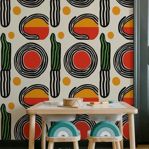 May include: A children's play area featuring a light wood table and two rainbow-shaped stools. The wall is decorated with a repeating pattern of black, red, yellow, and green abstract shapes. The floor is covered with a white rug.