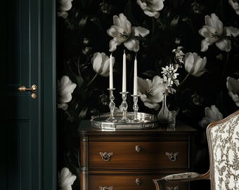 May include: A dark wood dresser with three drawers, topped with a silver tray holding three lit candles in crystal holders. The background features a black floral wallpaper with large white tulip blooms. A patterned chair is on the right.