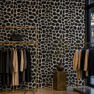 May include: A retail display featuring two gold metal clothing racks holding various garments, a black handbag, and a dark table with a black handbag and folded clothing. A dark vase with green branches sits on a gold pedestal against a patterned wall.