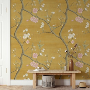 May include: Mustard yellow wallpaper featuring a floral design with branches, pink and white blossoms, and green leaves. A wooden bench, a woven basket, and a vase with cotton stems are in front of the wallpaper.