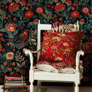 Op de afbeelding: Een witte stoel met een rood bloemenkussen en een patroon plaid. De achtergrond is een donker bloemenbehang met rode en oranje bloemen en groene bladeren. Een kleine bijzettafel houdt boeken vast.