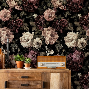 May include: A vintage wooden cabinet with a retro radio and potted plants sits in front of a floral wallpaper. The wallpaper features large, dark roses in shades of pink, cream, and burgundy against a black background. The cabinet is made of light brown wood.