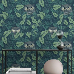 May include: Wallpaper with a repeating pattern of owls hidden amongst green leaves. The owls have detailed feathering and large eyes. The background is a deep teal, creating a contrast with the foliage and owl details. A shelf holds books, a vase, and an hourglass.