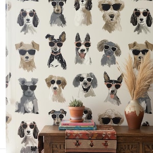 Puede incluir: Papel pintado con un patrón repetido de perros ilustrados con gafas de sol. Los perros son de varios colores y razas, sobre un fondo crema. Una mesa de madera con un jarrón, una planta y libros está frente al papel pintado.