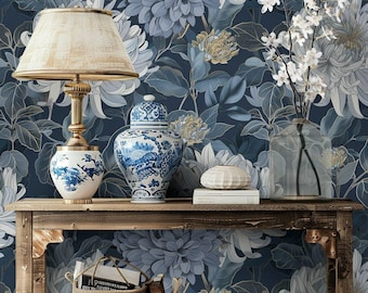 Puede incluir: Una consola de madera exhibe una lámpara, jarrones de porcelana azul y blanca y un jarrón de cristal con flores blancas. El fondo es un papel pintado floral en tonos azules y blancos, creando una estética elegante.