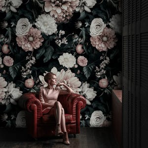 May include: A woman sits in a red leather armchair against a floral wallpaper backdrop. The wallpaper features large, stylized flowers in shades of white, pink, and peach against a dark background. The woman wears a light pink dress and brown boots.