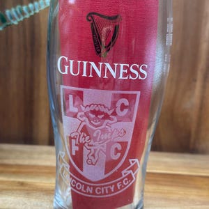 May include: A clear pint glass with the Guinness logo and a red background. The glass also features the text "Lincoln City F.C." and "The Imps" with a crest design. The glass is designed for beer.