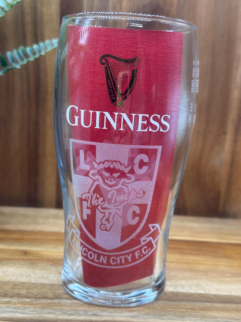 May include: A clear pint glass with the Guinness logo and a red background. The glass also features the text "Lincoln City F.C." and "The Imps" with a crest design. The glass is designed for beer.