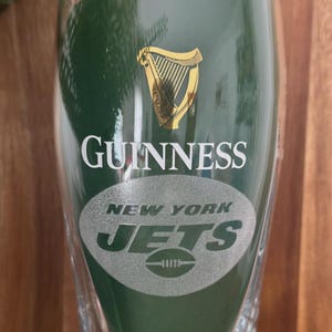 May include: A green Guinness pint glass with the Guinness logo and the New York Jets logo. The Guinness logo features a gold harp. The Jets logo is a silver football.