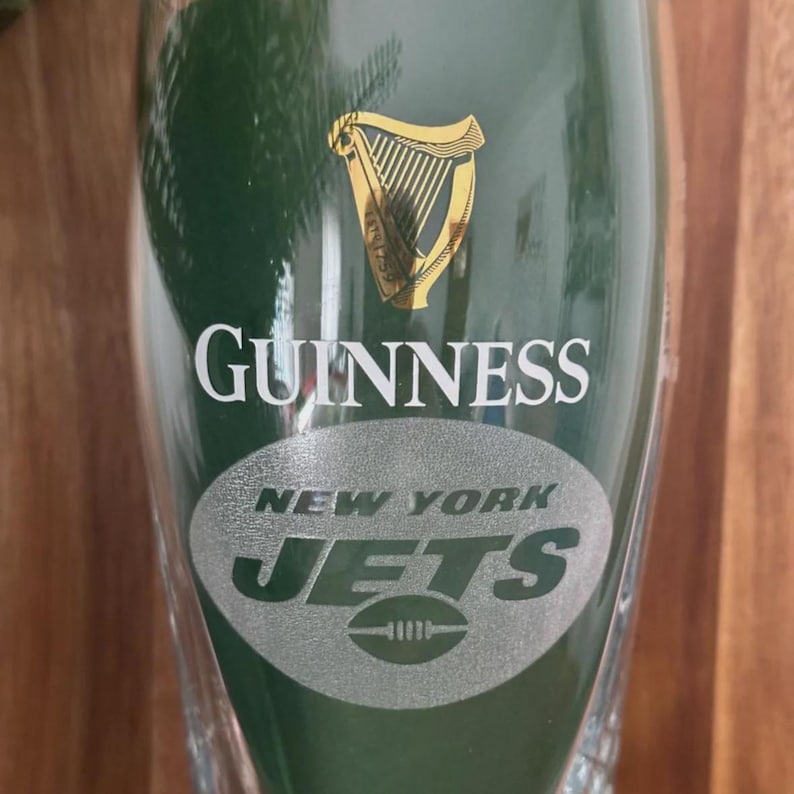 May include: A green Guinness pint glass with the Guinness logo and the New York Jets logo. The Guinness logo features a gold harp. The Jets logo is a silver football.