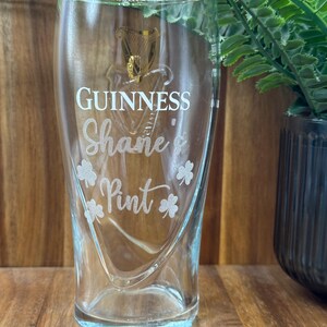 May include: A clear glass pint glass with the Guinness logo and the text "Shane's Pint" etched on the side. There are four shamrock designs around the text.