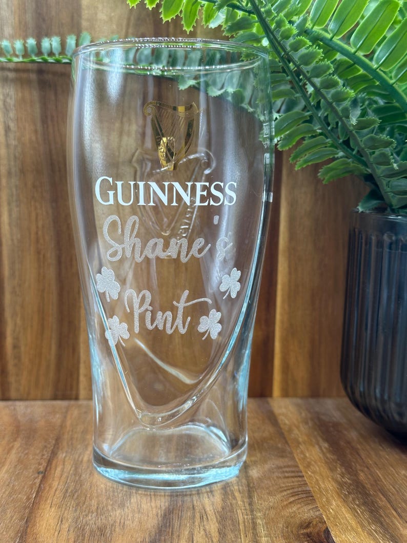 May include: A clear glass pint glass with the Guinness logo and the text "Shane's Pint" etched on the side. There are four shamrock designs around the text.