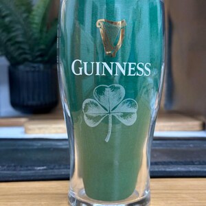 May include: A clear pint glass with the Guinness logo in gold and white. The glass features the Guinness name, a harp, and a shamrock design. The glass is filled with a green material, and is set against a blurred background.
