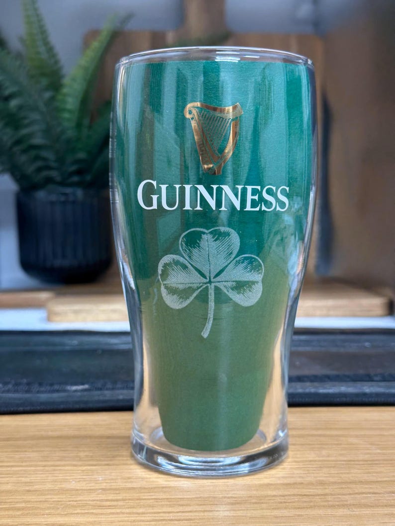 May include: A clear pint glass with the Guinness logo in gold and white. The glass features the Guinness name, a harp, and a shamrock design. The glass is filled with a green material, and is set against a blurred background.