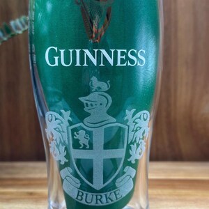 May include: A clear pint glass with a green background and the word "GUINNESS" in white. The glass features a crest with the name "BURKE" at the bottom. The Guinness harp logo is at the top.
