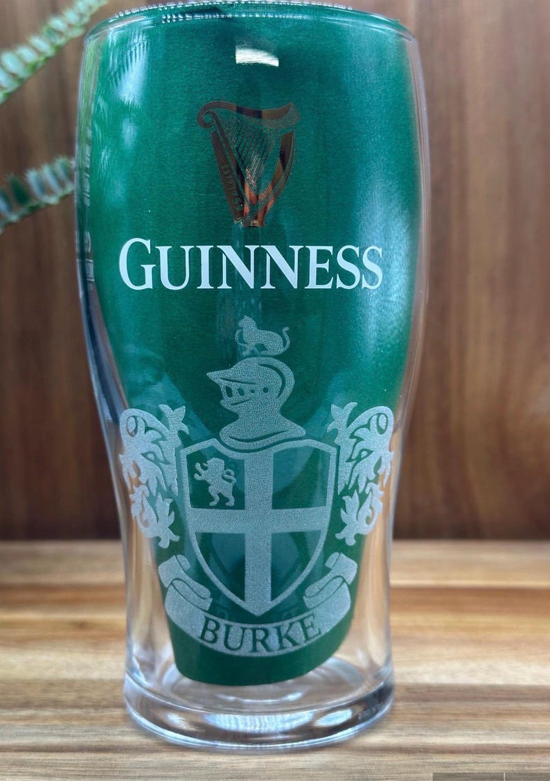May include: A clear pint glass with a green background and the word "GUINNESS" in white. The glass features a crest with the name "BURKE" at the bottom. The Guinness harp logo is at the top.