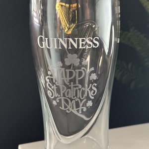 May include: A clear glass pint glass with a gold Guinness harp logo and the text "GUINNESS" etched on the front. The glass also has the text "Happy St. Patrick's Day" etched on the front with shamrock designs.
