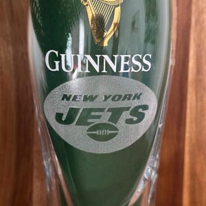 May include: A clear pint glass with a green background. The word "GUINNESS" is printed in white, with the gold harp logo above. Below, the words "NEW YORK JETS" and a football logo are etched in silver.