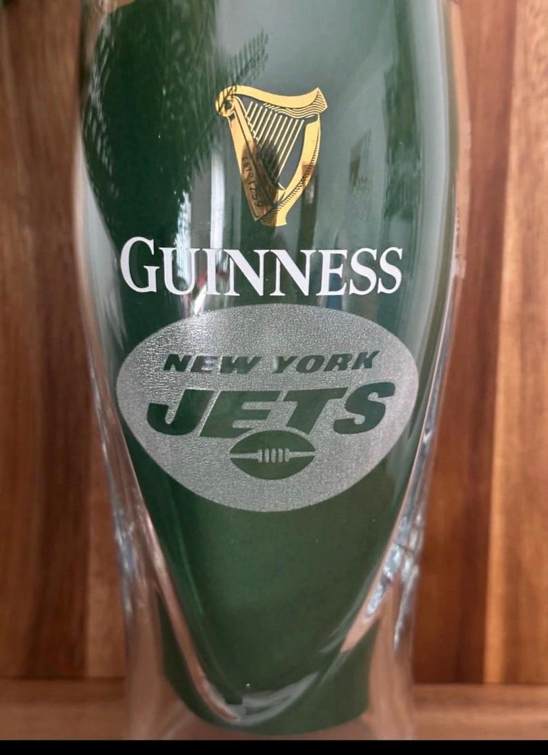 May include: A clear pint glass with a green background. The word "GUINNESS" is printed in white, with the gold harp logo above. Below, the words "NEW YORK JETS" and a football logo are etched in silver.