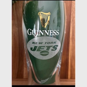 Op de afbeelding: Een groene Guinness pintglas met het New York Jets logo. Het glas heeft het Guinness logo in goud en het Jets logo in wit en groen. Het glas staat op een houten oppervlak.