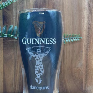 May include: A clear, double-walled pint glass with the Guinness logo, a harp, and the word "GUINNESS" in white. Below is a graphic of a person and the word "Harlequins". The glass is set against a wooden background.