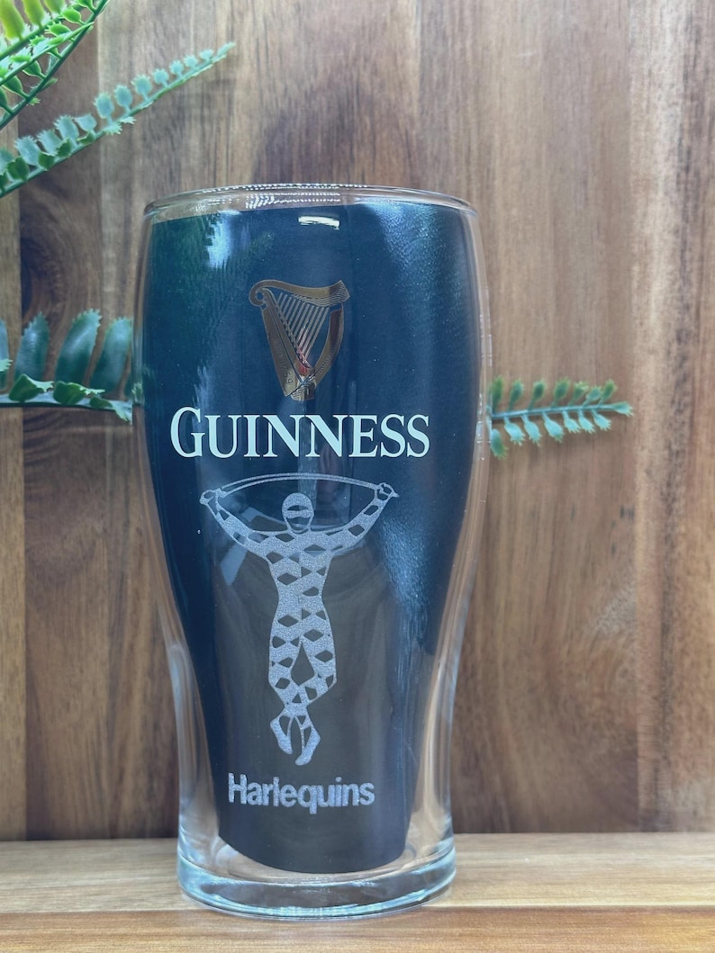 May include: A clear, double-walled pint glass with the Guinness logo, a harp, and the word "GUINNESS" in white. Below is a graphic of a person and the word "Harlequins". The glass is set against a wooden background.