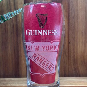 May include: A clear pint glass with a red background featuring the Guinness logo and the words "GUINNESS" in white. Below, the words "NEW YORK RANGERS" are displayed in a shield-shaped design.
