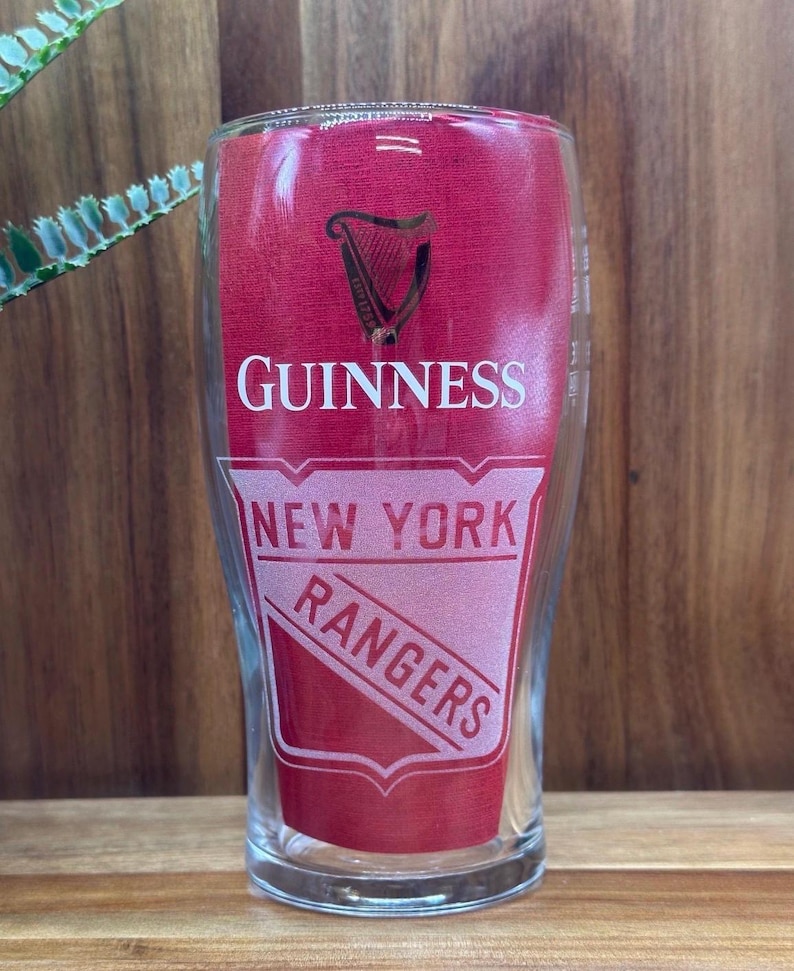 May include: A clear pint glass with a red background featuring the Guinness logo and the words "GUINNESS" in white. Below, the words "NEW YORK RANGERS" are displayed in a shield-shaped design.