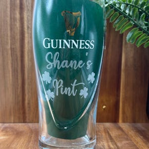 May include: A clear glass pint glass with a green background and gold Guinness logo. The glass is personalised with the text "Shane's Pint" and shamrock designs.