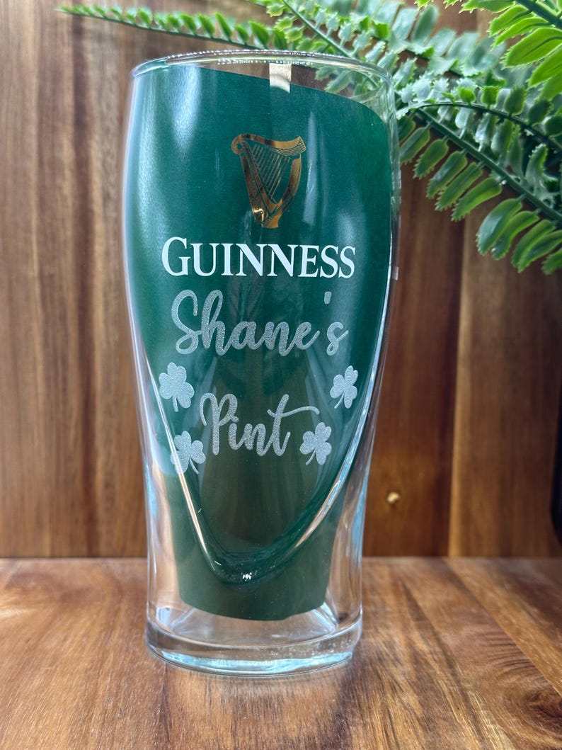 May include: A clear glass pint glass with a green background and gold Guinness logo. The glass is personalised with the text "Shane's Pint" and shamrock designs.