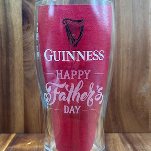 May include: A clear pint glass with a red insert featuring the Guinness logo and the words "Happy Father's Day" in white script. The glass has a curved shape and is designed for beer.